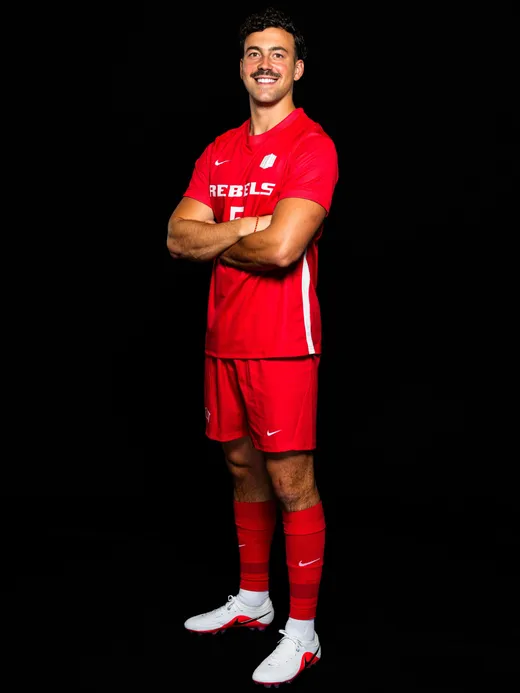 Manuel in the red UNLV Rebels kit, arms crossed and smiling, against a black background